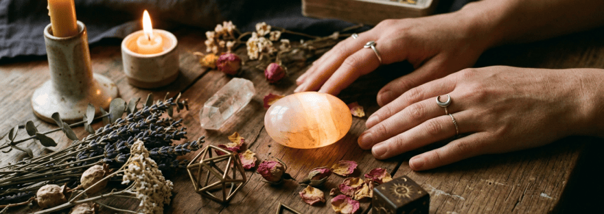 Hands beside glowing crystal, candles, dried flowers, and tarot cards